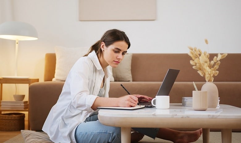 Eine junge Frau sitzt im Schneidersitz am Couchtisch, vor ihr ein Laptop. Sie schreibt sich Notizen in einen daneben liegenden Block.