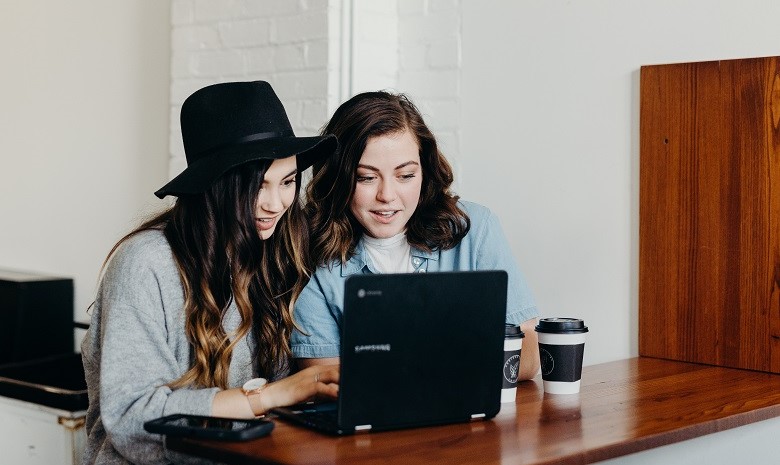Aktive Sprache Beitragsbild 1: Zwei junge Frauen sitzen am Tisch und schauen gemeinsam auf den Bildschirm eines Laptops