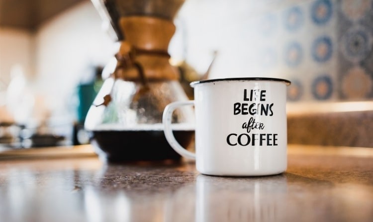 Eine weiße Tasse mit der Aufschrift "Life begins after Coffee" steht auf einer Anrichte vor einer Kanne Kaffee.