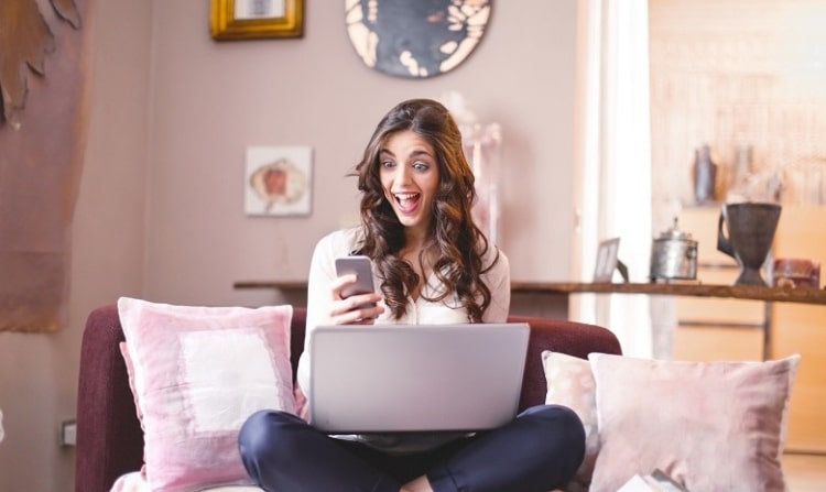 Werbetexte schreiben Headerbild: Eine junge Frau sitzt mit gekreuzten Beinen auf ihrem Sofa, den Laptop auf dem Schoss. In der rechten Hand hält sie ihr Smartphone, dessen Bildschirm sie begeistert ansieht.