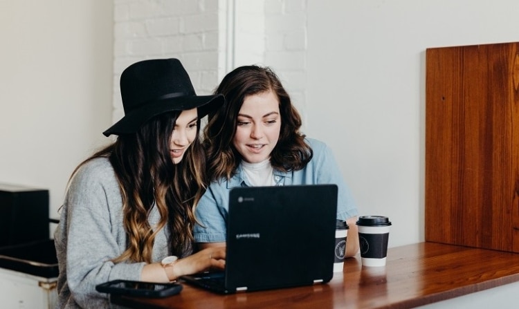Zwei junge Frauen schauen gespannt auf den Bildschirm eines Laptops. Auf dem Tisch daneben stehen zwei Becher Kaffee.